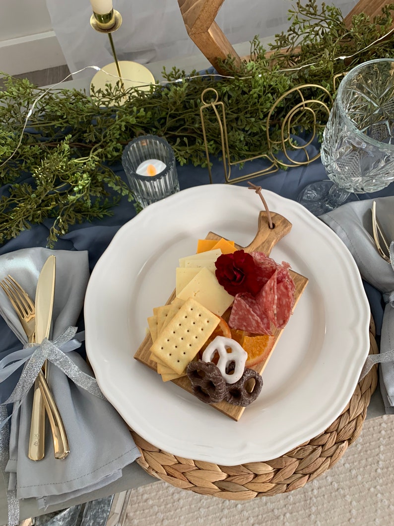 May include: A charcuterie board with crackers, cheese, salami, and pretzels on a wooden cutting board. The board is on a white plate with a scalloped edge. Gold silverware and a blue napkin are on the left. The word "love" is in gold wire.
