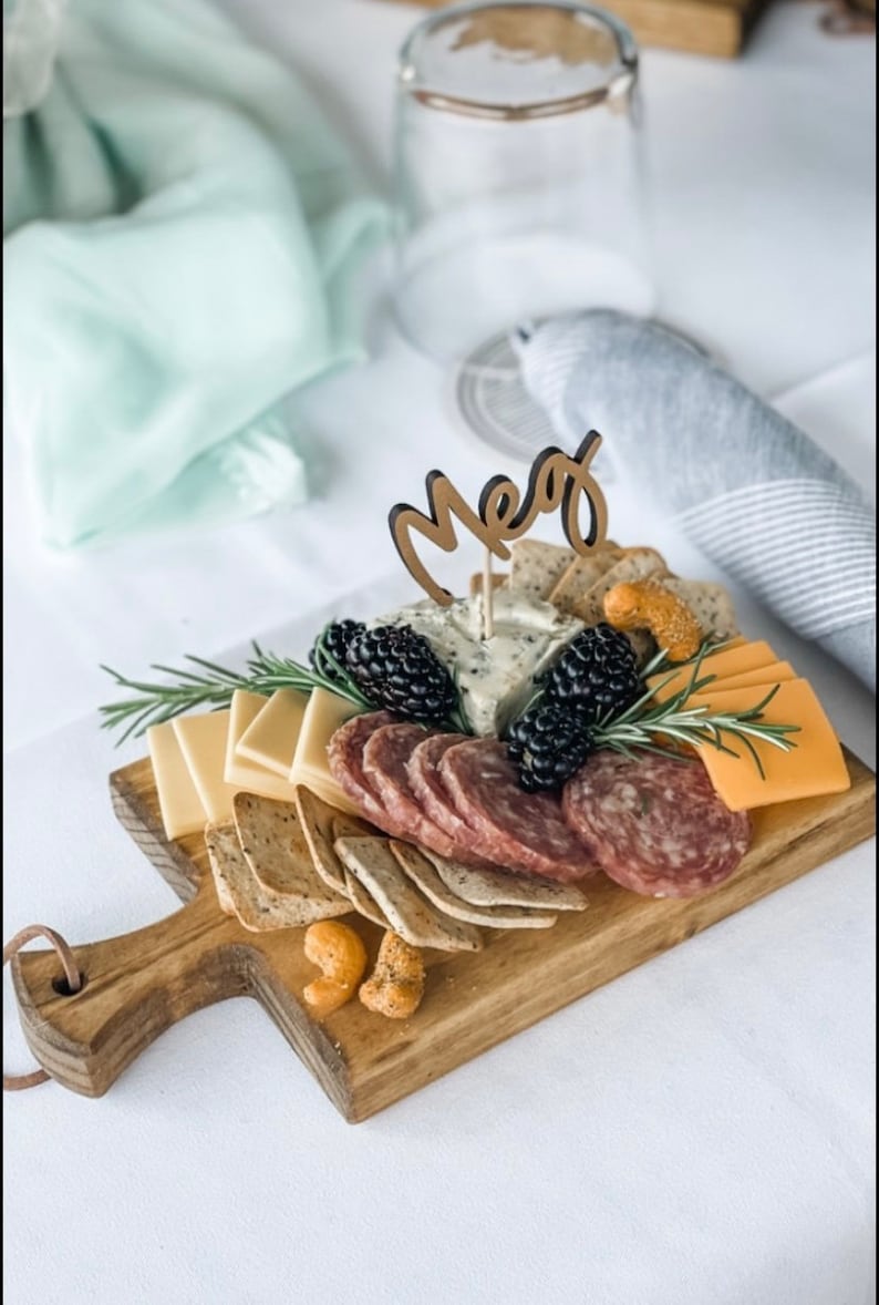 May include: Wooden charcuterie board with an assortment of cheeses, salami, crackers, and blackberries. The board is topped with a wooden "Meg" cake topper and sprigs of rosemary. A perfect appetizer for a gathering.