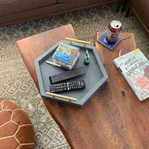 May include: A gray hexagonal tray with gold handles, two remote controls, a stack of postcards, and a tube of sunscreen. The tray is on a wooden table with a book titled "The World According to Fannie Davis" on the table.