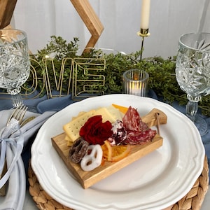 May include: A charcuterie board with crackers, cheese, salami, and dried fruit sits on a white plate. The plate rests on a woven placemat. A gold wire word "LOVE" and a candle are in the background.