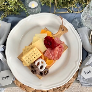 May include: A white ceramic plate with a wooden cutting board on top. The cutting board has a variety of snacks, including crackers, cheese, salami, fruit, and pretzels. The plate is on a woven placemat.
