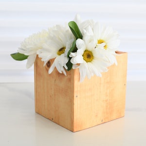 May include: A wooden square planter with white daisies in it. The planter is made of natural wood and has a light brown stain.