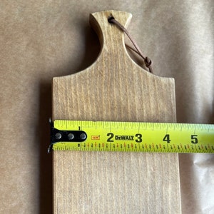 May include: A light brown wooden cutting board with a handle and a leather loop for hanging. The board is approximately 5 inches long, as measured by a yellow tape measure.