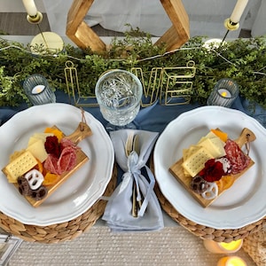 May include: A romantic picnic setting with two white plates holding charcuterie boards. Each board has crackers, cheese, salami, and fruit. Gold "LOVE" sign, candles, and a wooden geometric shape complete the scene. The table is set with blue linens.