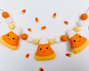 May include: A felt garland with orange, yellow, and white candy corn shapes. The candy corn shapes have smiling faces. The garland is strung with white, yellow, and orange felt balls.