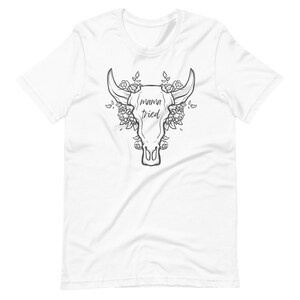 May include: White t-shirt featuring a black and white graphic of a bull skull with roses and the text "mama tried". The design is centered on the front of the shirt. The shirt has short sleeves and a crew neck.