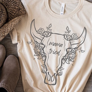 May include: Beige t-shirt with a graphic of a bull skull adorned with roses and the text "mama tried" in a handwritten style. A brown cowboy hat and boots are visible in the background.