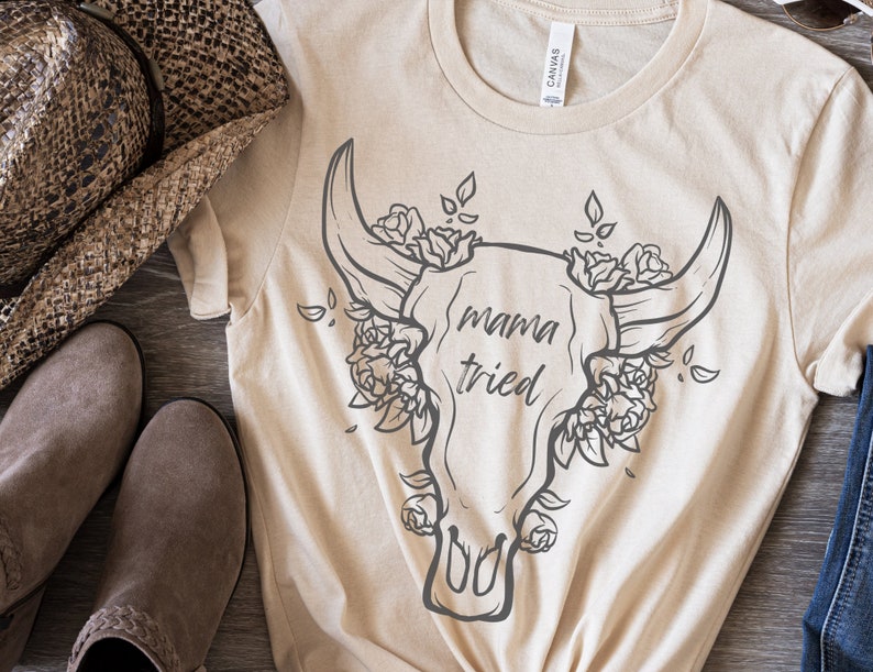 May include: Beige t-shirt with a graphic of a bull skull adorned with roses and the text "mama tried" in a handwritten style. A brown cowboy hat and boots are visible in the background.
