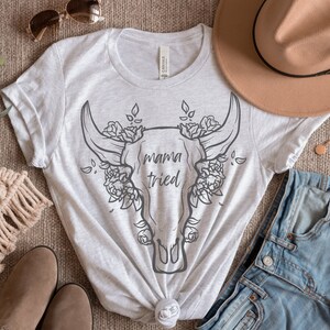 May include: A light gray t-shirt featuring a graphic of a bull skull with floral accents and the text "mama tried". The shirt is styled with a brown hat, denim shorts, and accessories.