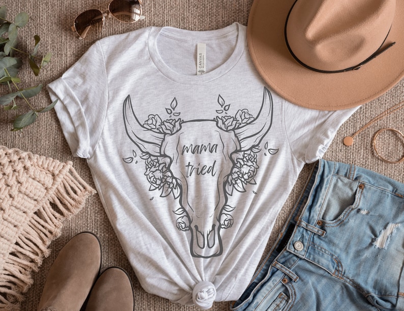 May include: A light gray t-shirt featuring a graphic of a bull skull with floral accents and the text "mama tried". The shirt is styled with a brown hat, denim shorts, and accessories.