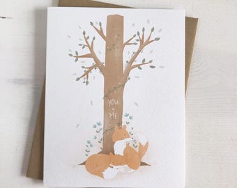 May include: A greeting card with a watercolor illustration of two foxes nestled at the base of a tree. The tree trunk has the words "You + Me" carved into it.