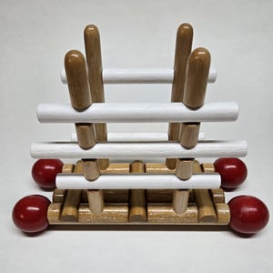 May include: A wooden puzzle toy with a light brown base and vertical posts. Horizontal white and light brown bars are connected to the posts. Red spheres are attached to the sides of the base. The toy is designed to challenge problem-solving skills.