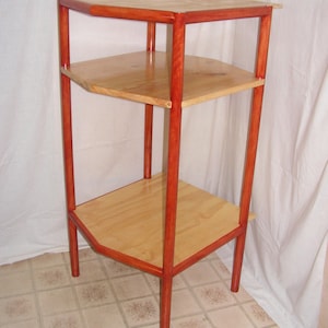 Puede incluir: Una estantería de madera de tres niveles con un acabado en madera natural y patas pintadas de rojo. Las estanterías son cuadradas y las patas son cilíndricas.