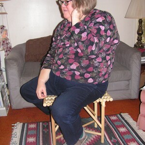 May include: A woman wearing glasses and a black and pink floral shirt sits on a wooden folding stool. The stool has a slatted seat and is on a rug with a geometric pattern.
