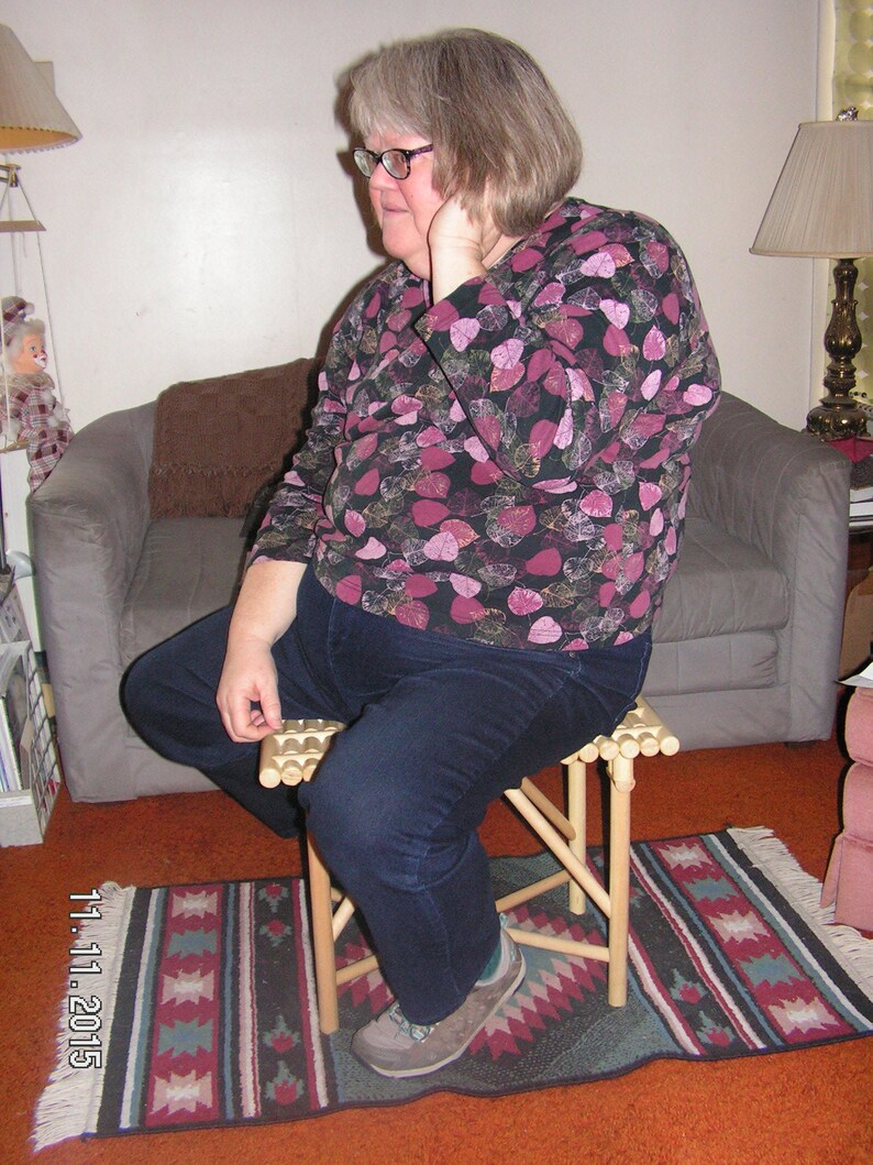 May include: A woman wearing glasses and a black and pink floral shirt sits on a wooden folding stool. The stool has a slatted seat and is on a rug with a geometric pattern.