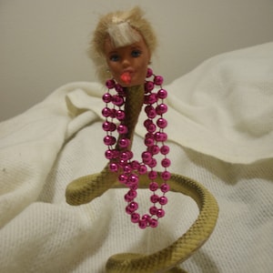 May include: A plastic doll head with blonde hair and pink lipstick is attached to a coiled, tan-colored snake. The doll is wearing a pink beaded necklace.