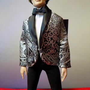 May include: A doll wearing a formal outfit. The doll is dressed in a black bow tie, a white dress shirt, a black and silver patterned blazer, and black pants. The blazer has a black shawl collar and a silver floral design.