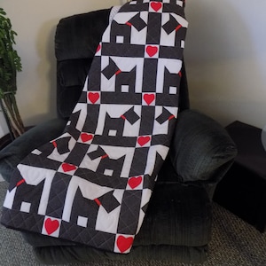 May include: A black and white quilt with a pattern of black silhouettes of dogs and red hearts. The quilt is folded and draped over a black armchair.
