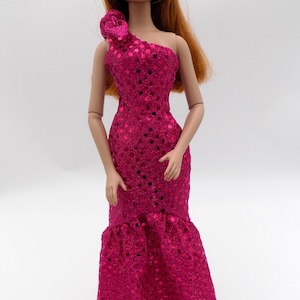 May include: A doll wearing a fuchsia, one-shoulder evening gown. The dress features a sequined bodice, a floral accent at the shoulder, and a flared hem. The doll has red hair and is wearing pearl earrings.