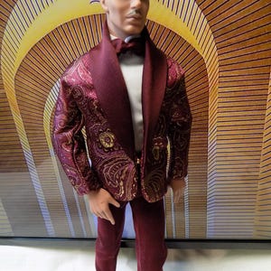May include: A doll wearing a burgundy suit with a paisley pattern and gold accents. The suit includes a jacket, pants, and a bow tie. The doll has a mustache and is set against a yellow and brown background.