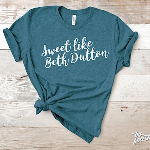 May include: Teal-colored t-shirt with the phrase "Sweet like Beth Dutton" in white script. The shirt is knotted at the bottom. The shirt is displayed on a light wood-grain surface.