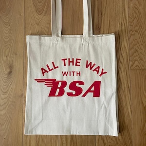 Pode incluir: Saco tote creme com texto vermelho "ALL THE WAY WITH BSA". O texto é em negrito, sem serifa. O saco tem duas alças e é feito de um material semelhante a lona.
