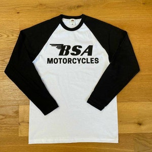 May include: White long-sleeve baseball t-shirt with black sleeves. The shirt has a black logo with a winged design and the text "BSA Motorcycles".