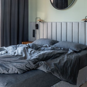Puede incluir: Un dormitorio moderno con una colcha y almohadas grises. Un espejo grande y redondo cuelga sobre la cama. El cabecero es de color gris claro y con paneles verticales. Cortinas grises oscuras enmarcan una ventana. Las paredes están pintadas de verde claro.