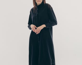 May include: A long-sleeved, dark navy blue shirt dress with a button-down front. The dress has a collared neckline and falls to mid-calf. The model is standing barefoot against a white background. The dress appears to be made of a soft, flowing fabric.