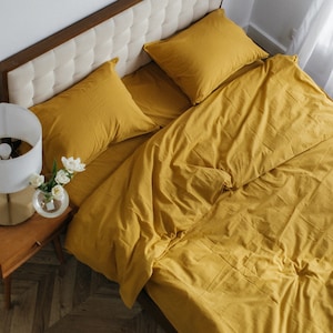 Peut inclure: Un lit avec une housse de couette jaune moutarde et deux oreillers assortis. La tête de lit est recouverte d'un tissu crème avec un cadre en bois. Une petite table de chevet avec une lampe et un vase de fleurs blanches est à côté du lit.