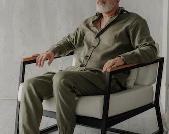 May include: A man sits in a black and white armchair with a wooden armrest. He is wearing a green linen shirt and matching trousers.