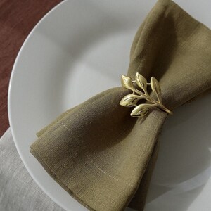 Puede incluir: Un anillo de servilleta de hoja de oro sobre una servilleta de lino marrón, doblada y colocada en un plato blanco.