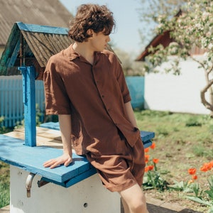 May include: A young person wearing a brown linen shirt and shorts sits on the edge of a well. The well is made of concrete and has a blue wooden top. The person is looking off to the side.
