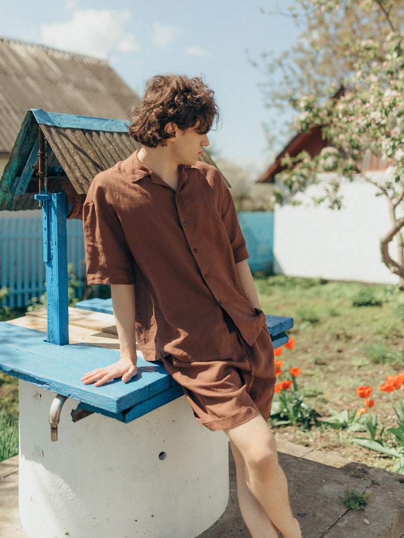 May include: A young person wearing a brown linen shirt and shorts sits on the edge of a well. The well is made of concrete and has a blue wooden top. The person is looking off to the side.