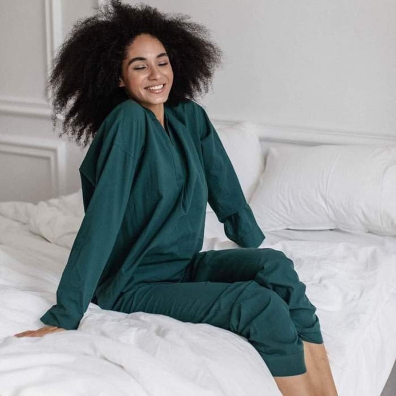 May include: A person wearing a dark green, long-sleeved pajama set with a V-neck top and matching pants. The person is sitting on a white bed with white pillows.