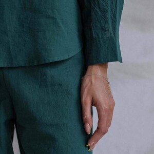 May include: A person wearing a dark green, long-sleeved shirt and matching pants. The shirt has a button-down collar and the pants have a straight leg. The person is wearing a silver chain bracelet on their wrist.