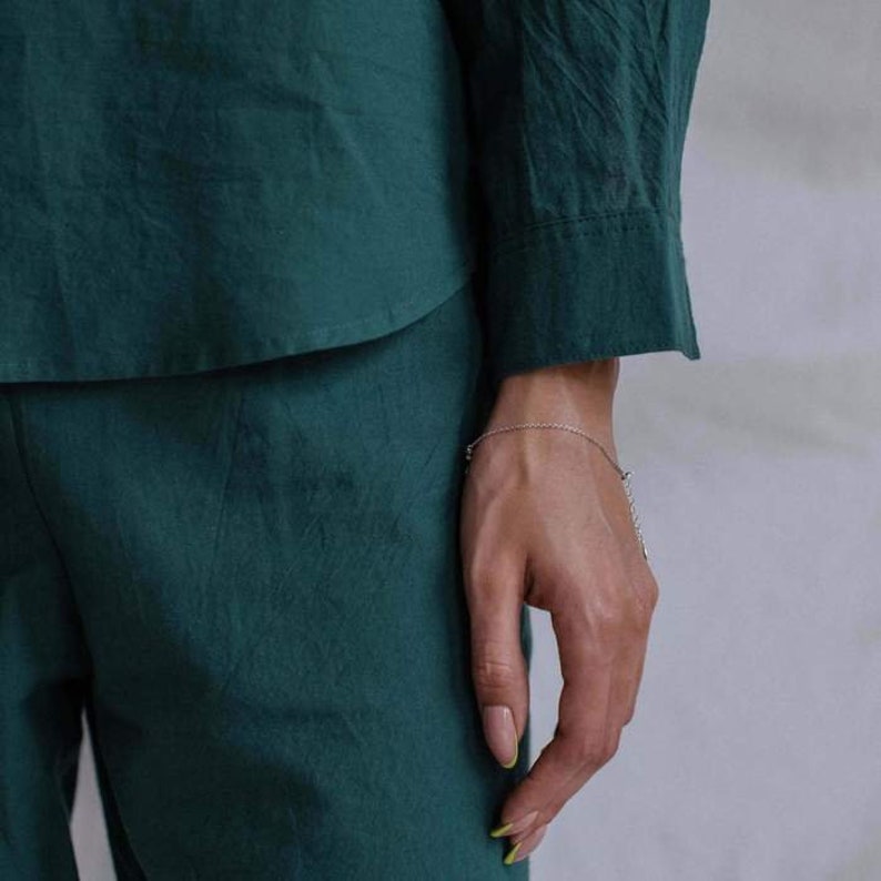 May include: A person wearing a dark green, long-sleeved shirt and matching pants. The shirt has a button-down collar and the pants have a straight leg. The person is wearing a silver chain bracelet on their wrist.