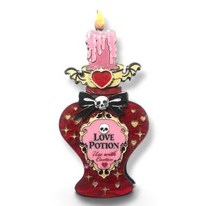 May include: A heart-shaped enamel pin featuring a red bottle with a pink candle on top. The bottle has a black bow with a skull, and a label that reads LOVE POTION Use with Caution. The design includes gold star and heart accents and bat wings.