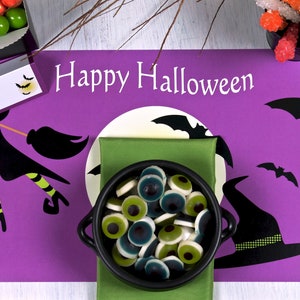 Puede incluir: Mantel individual de Halloween morado con una bruja volando en una escoba, murciélagos, una luna y el texto "Happy Halloween". Un caldero negro lleno de caramelos de ojos verdes y azules se encuentra sobre una servilleta verde.