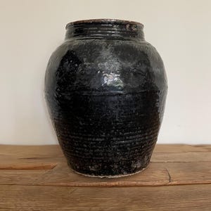 May include: A large, dark-glazed ceramic jar with a wide body and a slightly flared rim. The jar has a textured surface, with horizontal ridges around the lower portion. The color is a deep, glossy black, suggesting an antique or vintage piece.