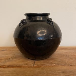 May include: A large, round, black ceramic vase with a glossy finish. The vase has a wide, bulbous body and a narrower neck with two small handles. The lower portion of the vase is a darker shade of black than the upper portion.