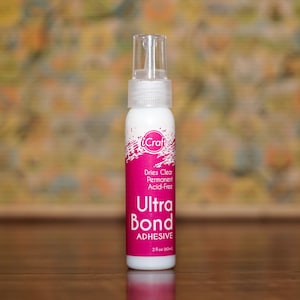 May include: A white bottle of iCraft Ultra Bond Adhesive with a clear spray nozzle. The label features pink accents and text that reads "Dries Clear, Permanent, Acid-Free." The bottle contains 2 fl oz (59 ml) of adhesive.