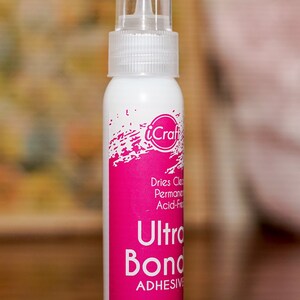 May include: A white bottle of iCraft Ultra Bond adhesive with a clear cap. The bottle has a bright pink label with white text that reads "Ultra Bond Adhesive" and "2 fl oz (60 ml)". The adhesive is acid-free and dries clear.