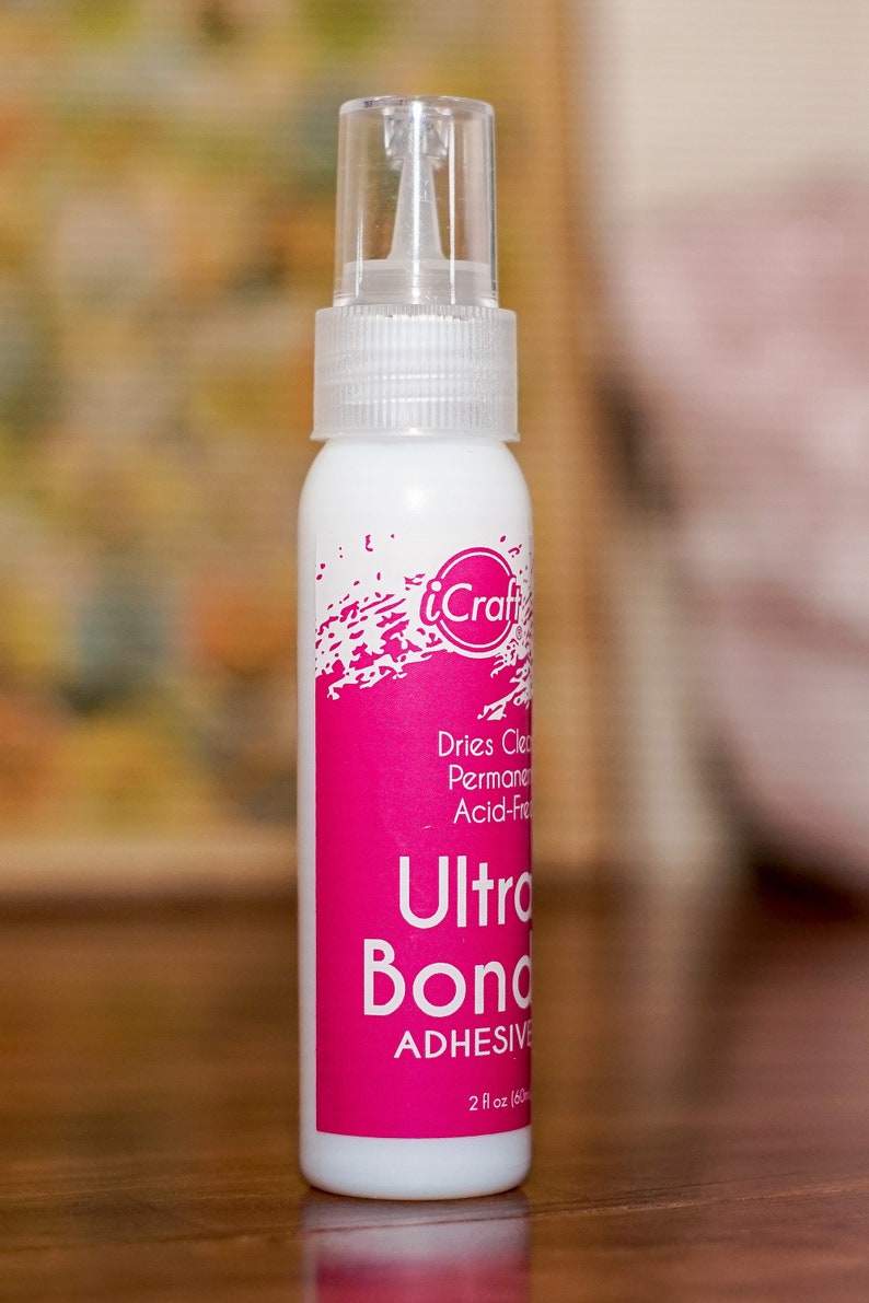 May include: A white bottle of iCraft Ultra Bond adhesive with a clear cap. The bottle has a bright pink label with white text that reads "Ultra Bond Adhesive" and "2 fl oz (60 ml)". The adhesive is acid-free and dries clear.