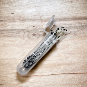 May include: A clear plastic tube filled with black headed pins. The tube has a white lid and is open, revealing the pins.