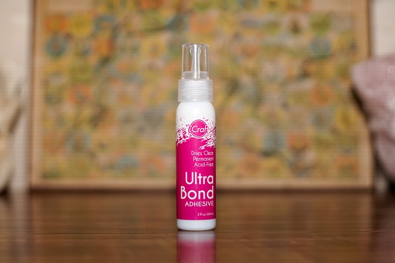 May include: A white bottle of iCraft Ultra Bond Adhesive with a clear spray nozzle. The label features pink accents and text that reads "Dries Clear, Permanent, Acid-Free." The bottle contains 2 fl oz (59 ml) of adhesive.