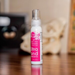 May include: A white bottle of iCraft Ultra Bond adhesive with a clear spray nozzle. The label is pink and white, with the text "Dries Clear Permanent Acid-Free". The bottle contains 2 fl oz (60ml) of adhesive.