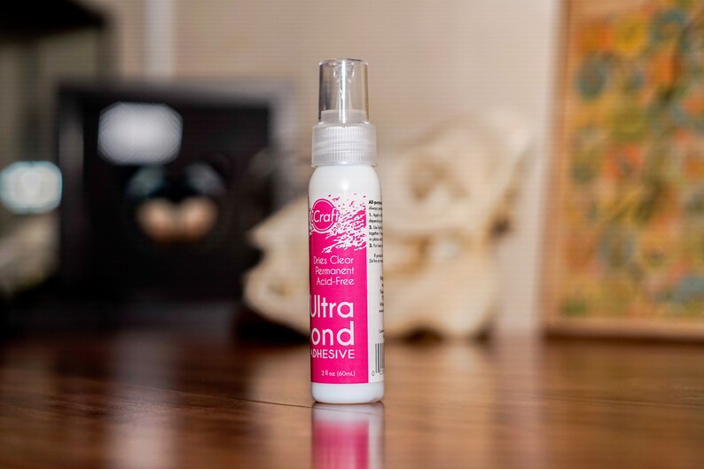 May include: A white bottle of iCraft Ultra Bond adhesive with a clear spray nozzle. The label is pink and white, with the text "Dries Clear Permanent Acid-Free". The bottle contains 2 fl oz (60ml) of adhesive.