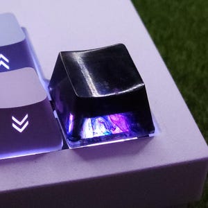May include: Close-up of a white mechanical keyboard with purple keycaps. The keycaps have white lettering and symbols. One keycap is a clear, glossy black with purple and blue internal lighting. The keyboard sits on a green surface.