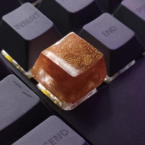 May include: Close-up of a mechanical keyboard with custom keycaps. The keycaps are black, with a few custom keys in clear and brown resin with gold glitter. The keyboard has a green background.